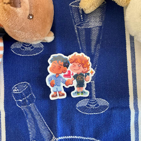 Foto de la pegatina hollanov de la serie Heated Rivalry. A modo de decoración a su alrededor hay puesto un mantelito azul y un peluche de un osito marrón.