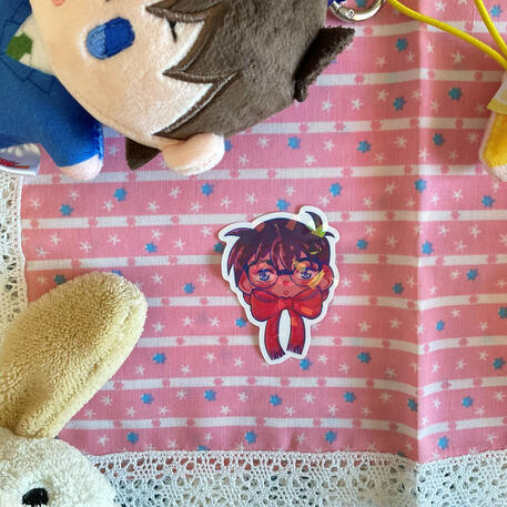 Foto de la pegatina de Conan del manga Detective Conan. A modo de decoración a su alrededor hay: un peluche de un perro blanco y un peluche de Shinichi del manga Detective Conan.