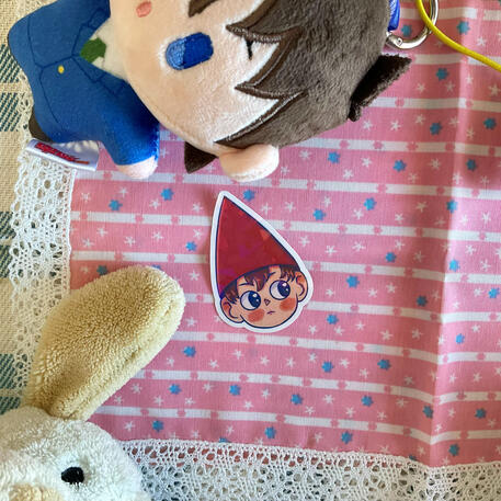 Foto de la pegatina de Wirt de la serie Over the Garden Wall. A modo de decoración a su alrededor hay: un peluche de un perro blanco y un peluche de Shinichi del manga Detective Conan.
