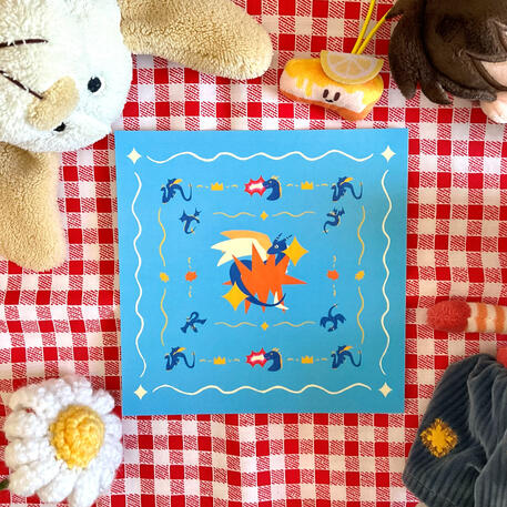 Foto de la print de un dragón azul. A modo de decoración a su alrededor hay: un peluche de un perrito, una flor de crochet blanca y un peluche de una tarta con cara.