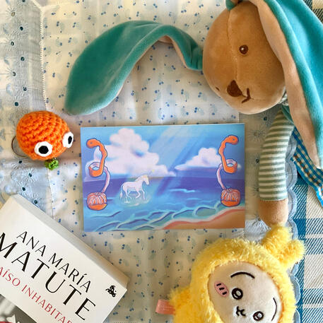 Foto de la print de un unicornio sobre el mar. A modo de decoración a su alrededor hay: una naranja con cara de crochet, el libro "Paraíso inhabitado" de Ana María Matute, un peluche de un conejito azul y un peluche de Usagi de la serie Chiikawa.